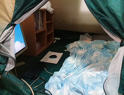 テントの中の写真。布団が敷いてあり、キーボードとマウスとモニターがある。奥には本棚がある。
